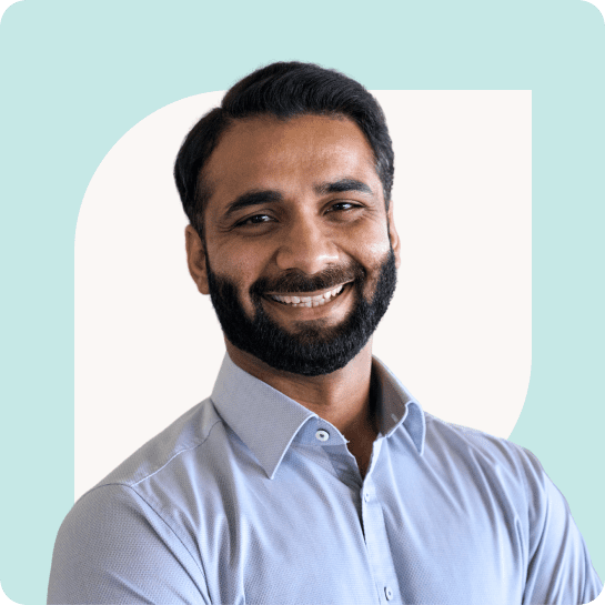 A man with a beard smiling at the camera, wearing a light blue shirt, standing against a light teal background.