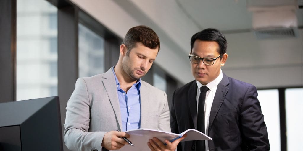 Two men in business attire are reviewing documents together in an office setting with large windows in the background.