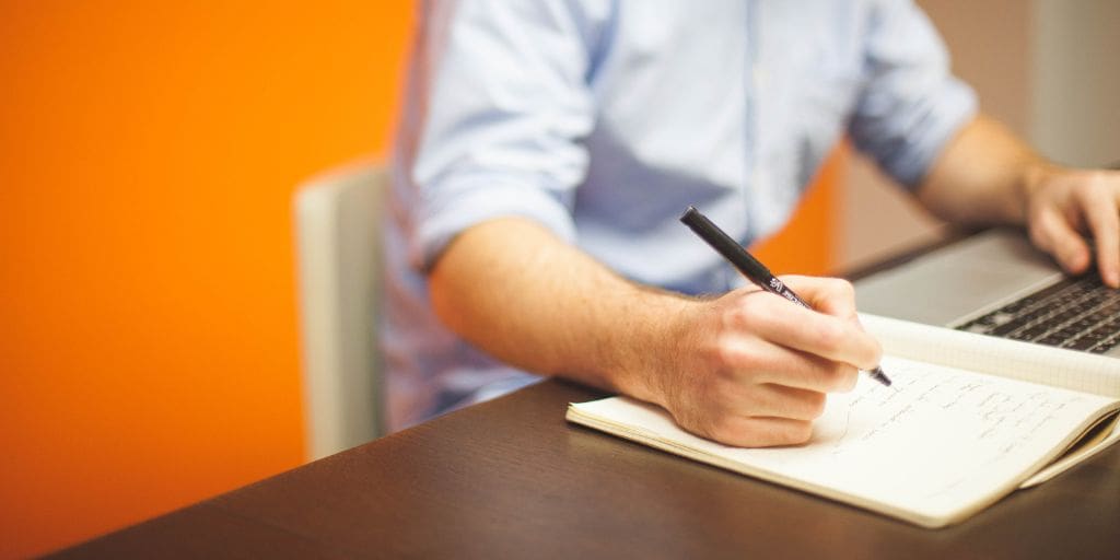 A person writes in a notebook with a pen while working on a laptop at a desk. An orange wall is visible in the background.