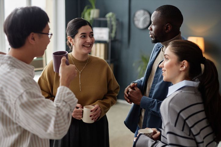 Multiethnic group of four cheerful adults as colleagues chatting and enjoying coffee break in office